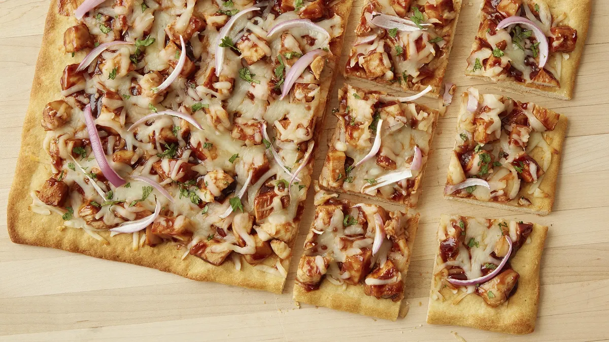 A flatbread pizza topped with barbecue chicken, melted cheese, red onion slices, and chopped herbs, cut into square pieces.