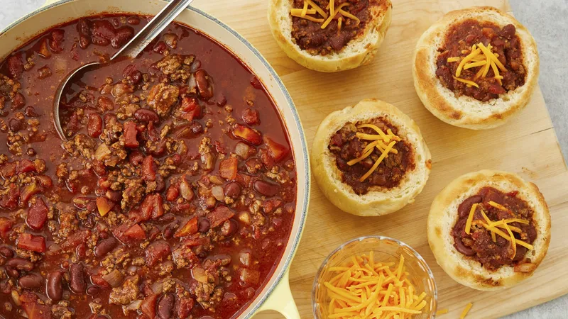 Large dutch oven with chili on top of wooden board with biscuit cups filled with chili and topped with shredded cheese.