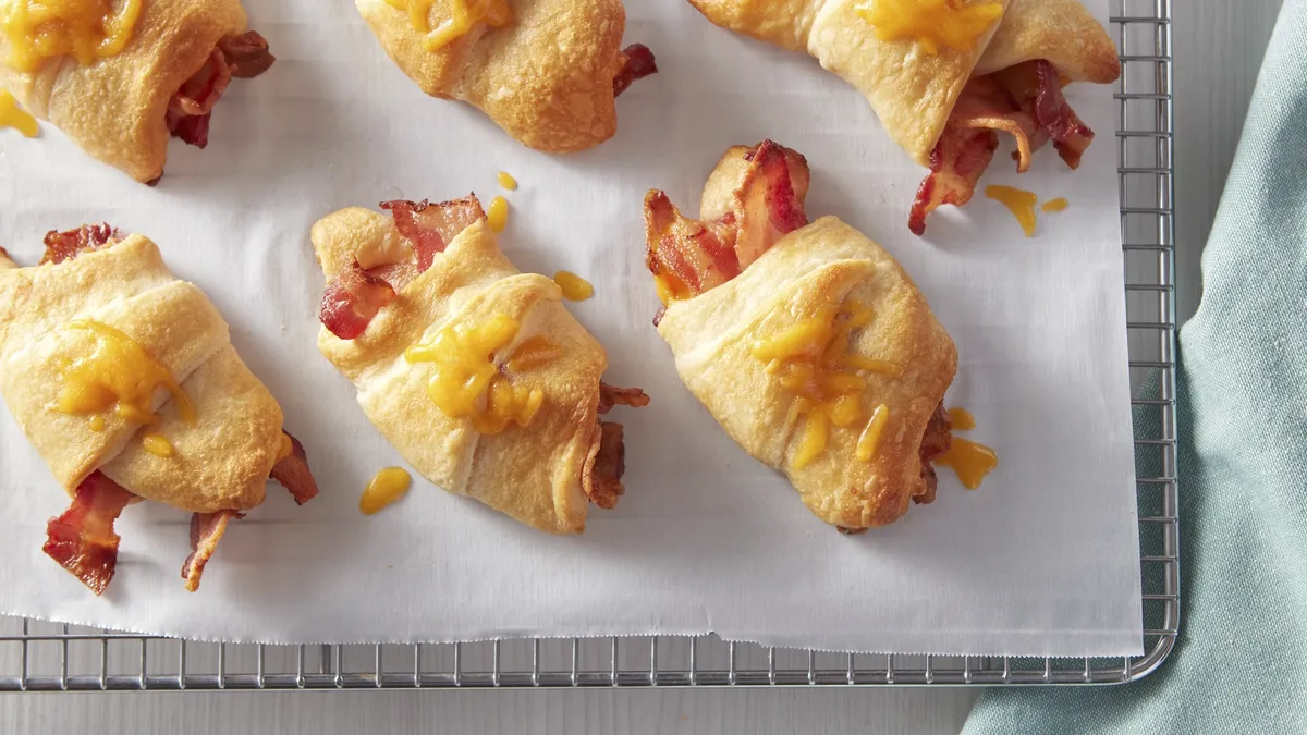 Baked crescent roll sandwiches filled with bacon and melted cheese on a parchment-lined cooling rack.