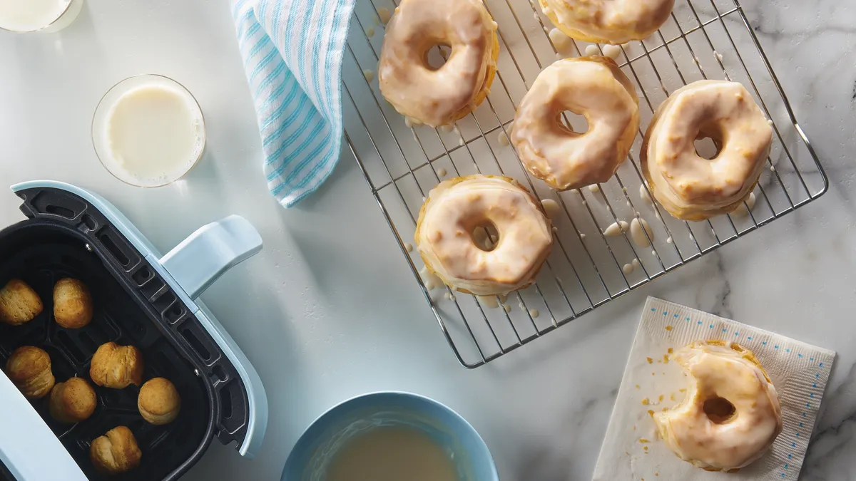 Freshly glazed doughnuts cooling on a wire rack with doughnut holes frying in an air fryer nearby.