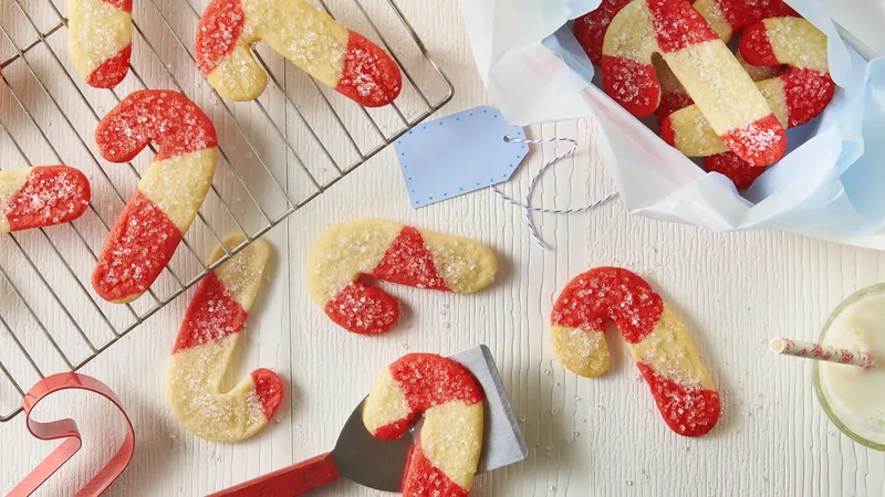 Top view of 4-Ingredient Candy Cane Cookies on a cooling rack and white table. The cookies are shaped like candy canes with red and white striped dough made from Pillsbury Sugar Cookie Dough, topped with sugar. A spatula, a blue napkin, and a glass of milk with a candy cane are also visible.