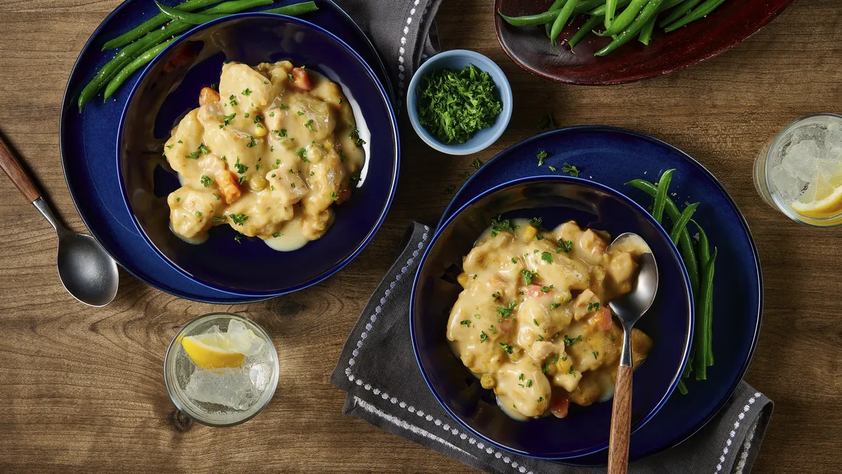 Two plates of creamy stew with vegetables, garnished with herbs, beside green beans and lemon water on a wooden table.