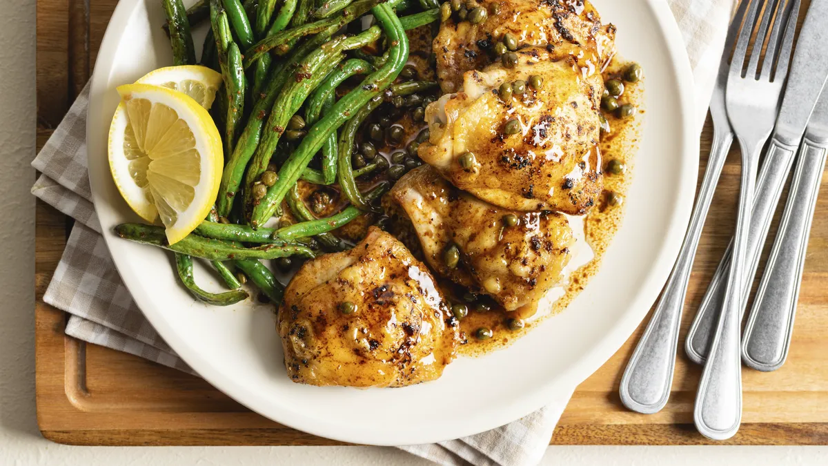 A photo of golden-brown chicken thighs in a pan sauce with capers, served on a white plate with roasted green beans and lemon slices.