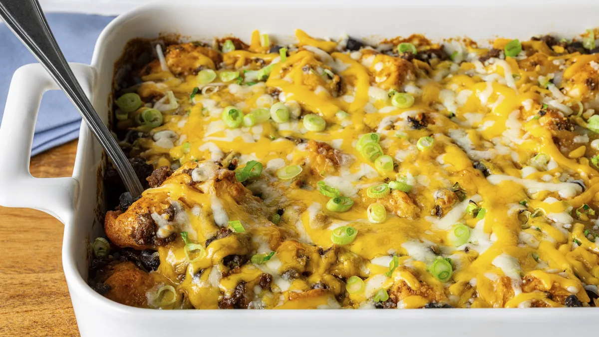 A casserole dish featuring puffy, bite-sized biscuit pieces mixed with seasoned ground beef, black beans, enchilada sauce, and melted Mexican-style cheese, garnished with fresh sliced green onions.