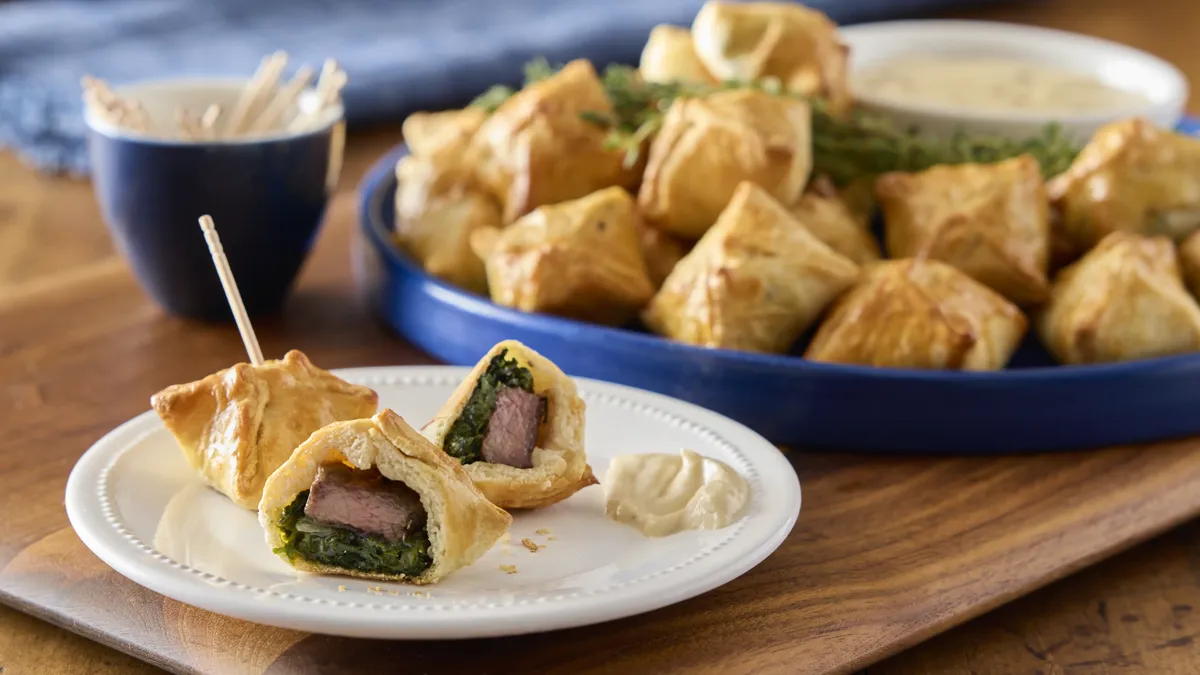 Several mini beef Wellingtons arranged on a serving tray are garnished with fresh thyme. The Wellingtons consist of golden-brown, flaky crescent dough folded over seared beef tenderloin and a creamy spinach mixture, and are served with a smoked gouda dipping sauce in a small white bowl.