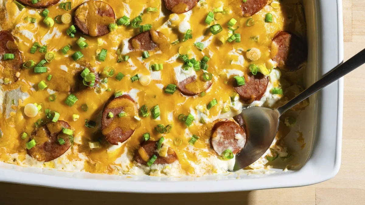 White casserole dish with pierogi, kielbasa, and a creamy cheese sauce topped with green onions. The dish is on top of a serving towel.