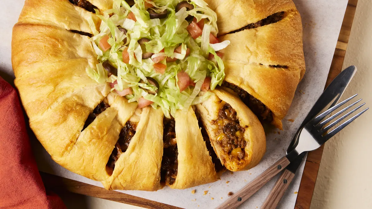 A crescent ring filled with seasoned taco meat and melted cheese. The center of the ring is filled with fresh shredded lettuce and chopped red tomatoes. To the right of the ring there is a fork and a knife. There is a red towel to the left side of the ring.