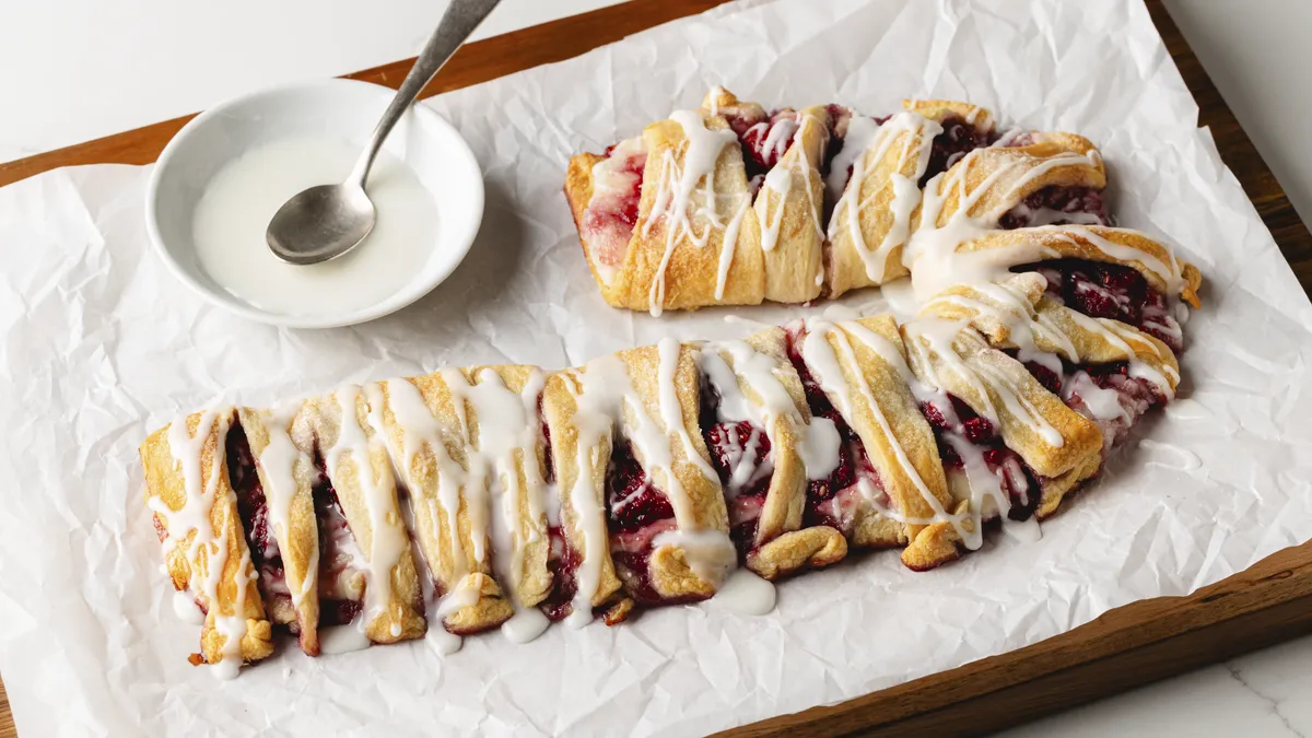 A holiday raspberry cream cheese crescent Danish features a flaky, braided golden crust with red raspberry filling and white icing drizzle, baked in the shape of a candy cane.