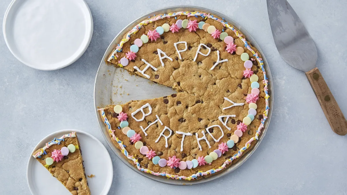 A giant cookie on a serving platter with the words “HAPPY BIRTHDAY” written on it in white icing surrounded by pastel candy sprinkles. A slice of the cookie is on a white plate and other white plates for serving are stacked near it. A pastry server is next to the giant cookie.