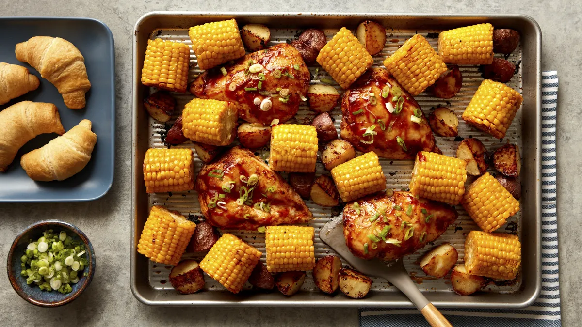 A parchment-lined baking sheet containing glazed chicken breasts, roasted potatoes, and cor with a spatula scooping up a chicken breast. To the left of the baking sheet there is a blue plate with golden-brown crescent rolls. Next to that plate there is a small bowl of green onions for garnishing.