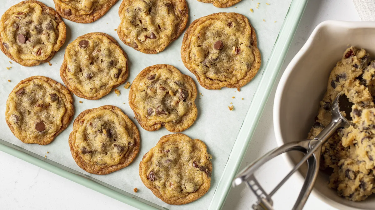 Bowl of chocolate chip cookie dough with a scoop alongside a baking sheet of baked cookies.