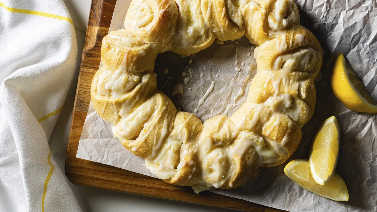 A photo of a golden-brown crescent ring drizzled with white icing on a parchment-lined wooden cutting board, with a few lemon slices sitting next to it.