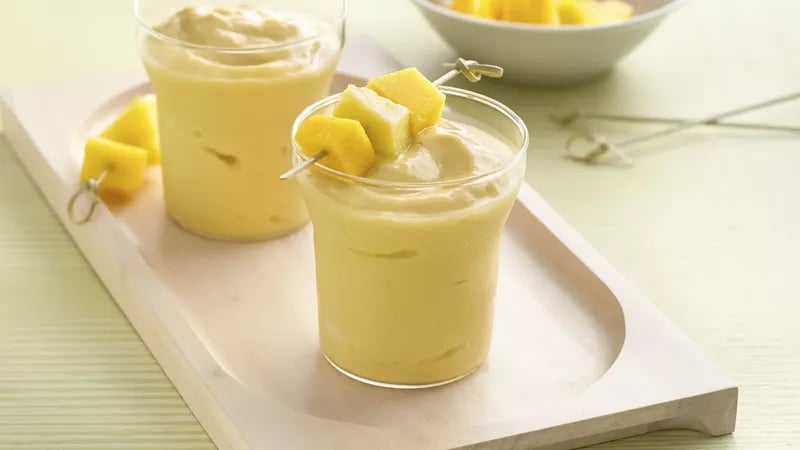 Two glasses of creamy mango smoothies garnished with mango chunks on skewers, served on a wooden tray.