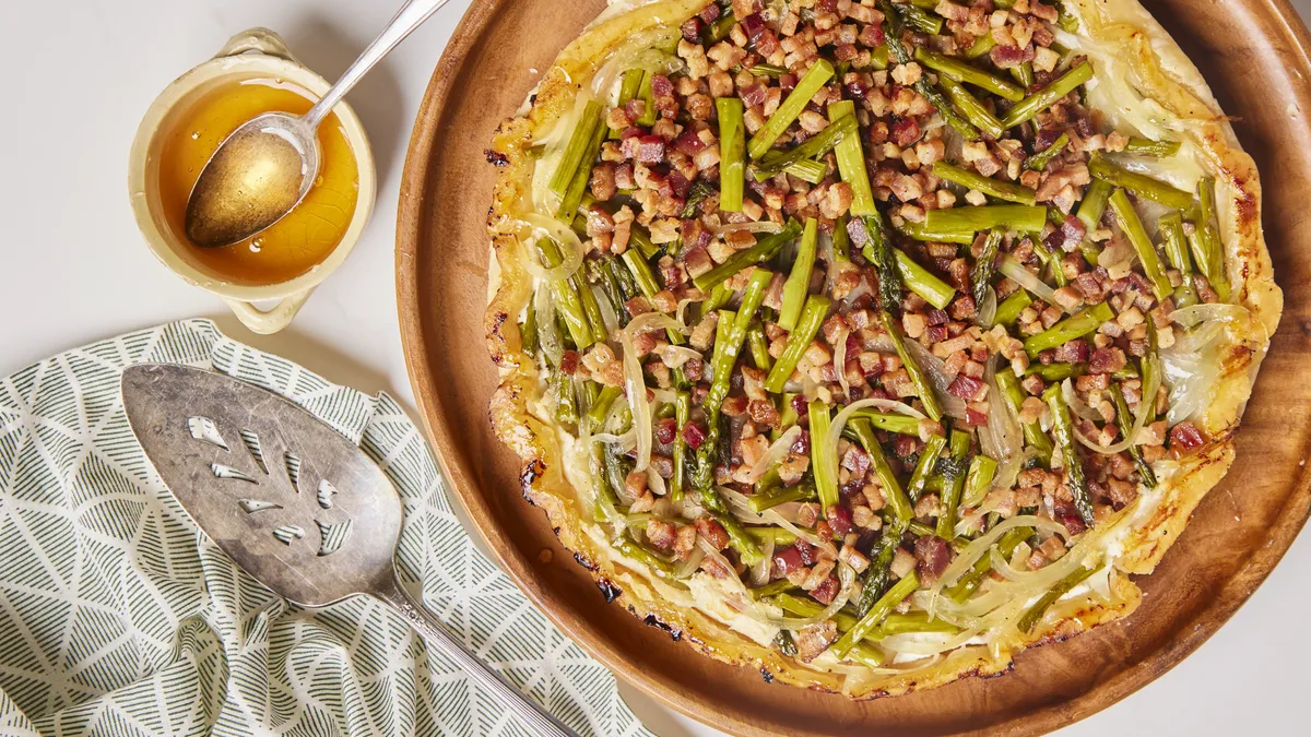 A circular savory tart with a rustic, golden-brown pie crust base. The top is covered in a colorful arrangement of vibrant green asparagus spears, golden caramelized onions, and bits of crispy bacon, all lightly glistening with olive oil and melted cheese.