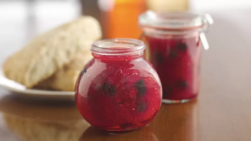 Jars of mixed berry jam next to a plate of biscuits on a wooden table.