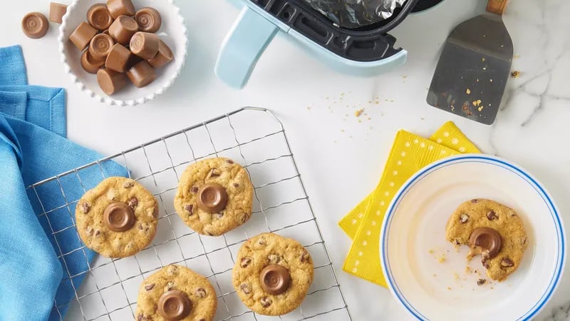 Top view of Air Fryer Caramel Chocolate Chip Cookies on a cooling rack and white table. The cookies, made from Pillsbury chocolate chip cookie dough, are topped with melty caramel-chocolate candies. An air fryer, a blue napkin, a yellow napkin, a plate with a half-eaten cookie, and a bowl of Rolo's® are also visible.