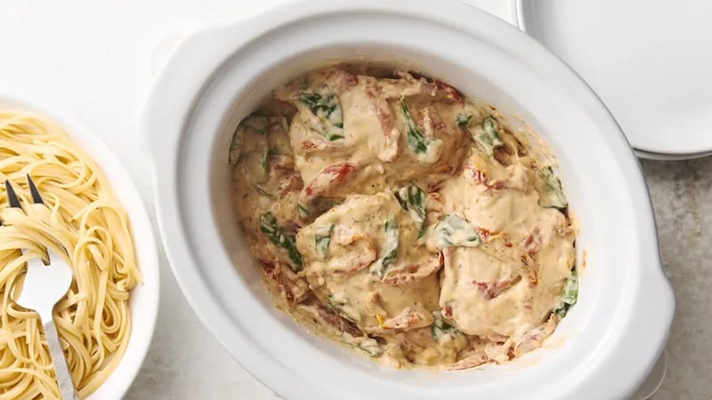 Top view of Slow-Cooker Creamy Tuscan Chicken in a white slow cooker. The dish features chicken, Alfredo sauce, sun-dried tomatoes, spinach, and Parmesan cheese. Next to the slow cooker is a white plate of cooked pasta, ready to be served