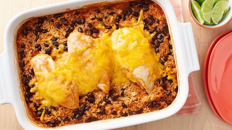 Casserole dish with rice, beans, chicken breasts, enchilada sauce and melted cheese on a peach cloth napkin, alongside a dish of lime wedges and stacked peach colored plates.